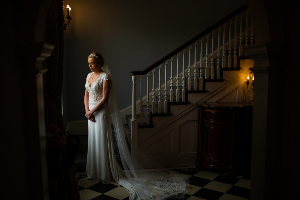 A bride looking down smiling as she stands near a large window at the bottom of a staircase