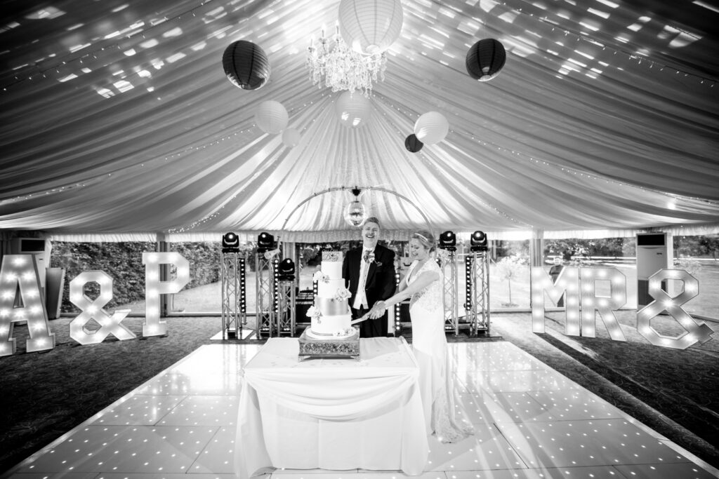 A black and white photo of a bride and groom cutting their cake on the dance floor in a marquee