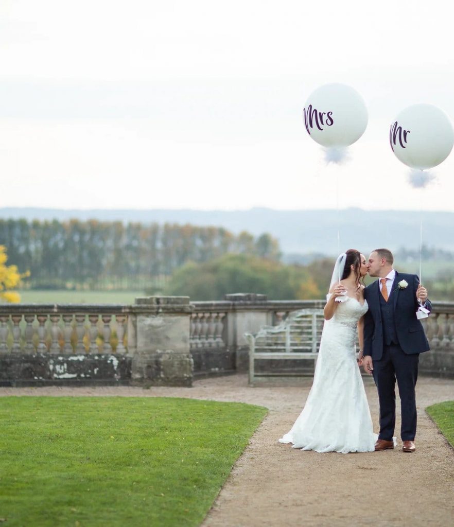 Bride and groom kiss whilst holding large Mr and Mrs balloons