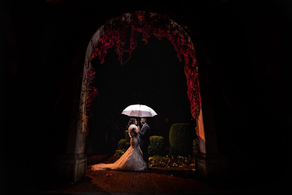 Bride ands Groom standing under a white umbrella in an archway lit up in red