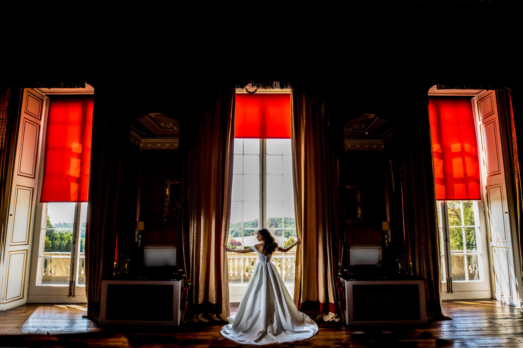 A bride holding curtains in a large stately home reception room. The curtains a red and the sun is shining in from the windows