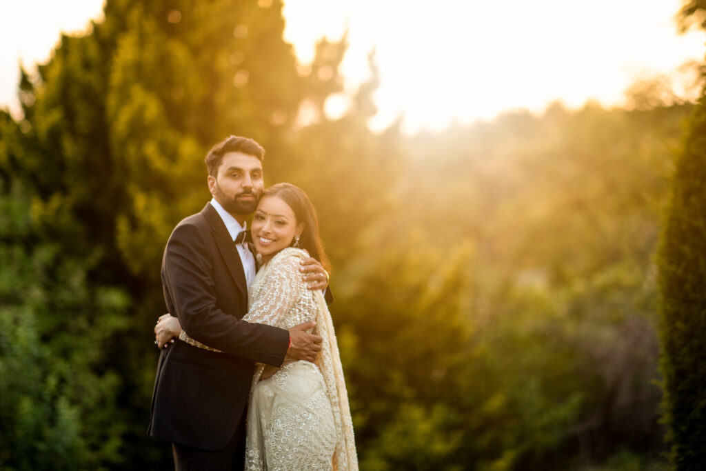 Bride and groom huggin each other smiling at the camera as the sun is setting behind them in a countryside side backdrop