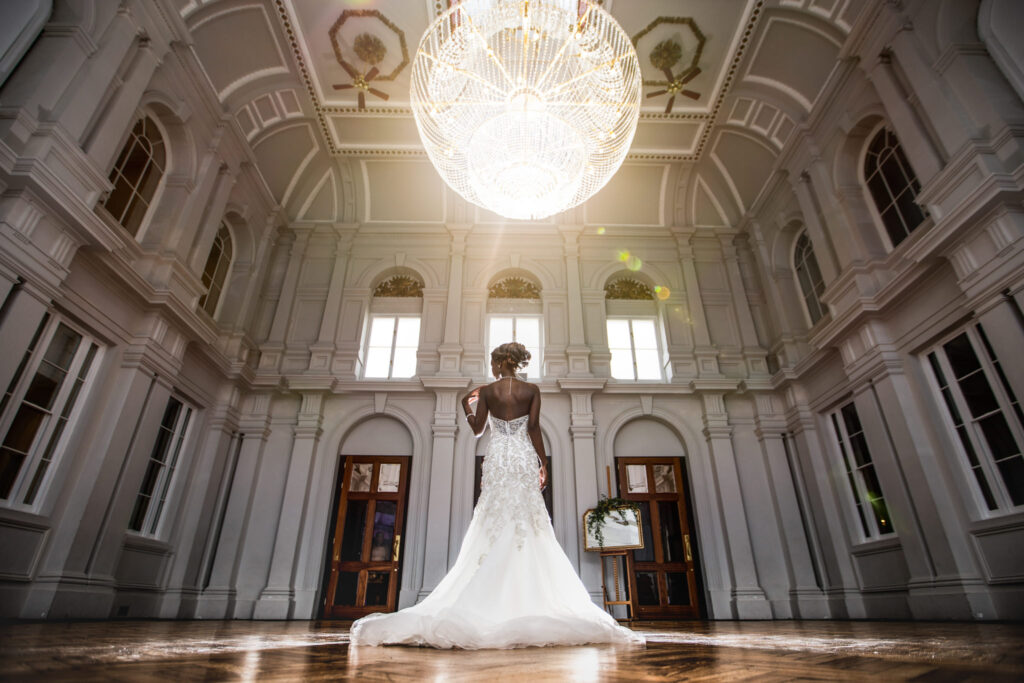 Bride standing away from the camera showing the back of her dress. She is standing underneath a large chandelier in a hall reception area.