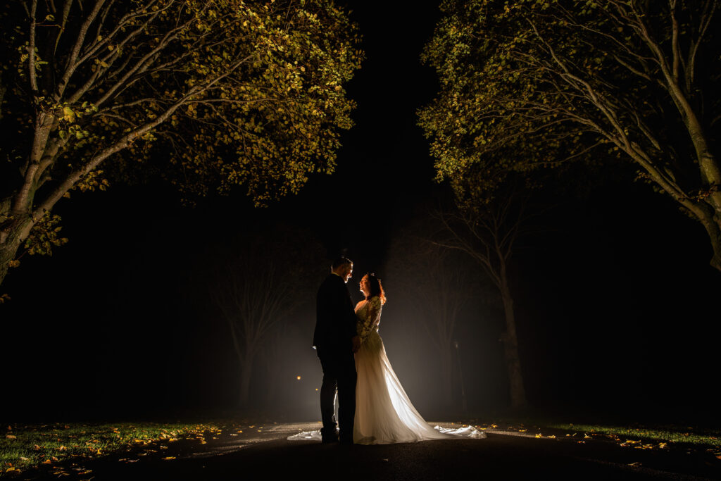 Bride and Groom standing looking at each other as they are lit by a bright light behind them. They are standing underneath two large trees.
