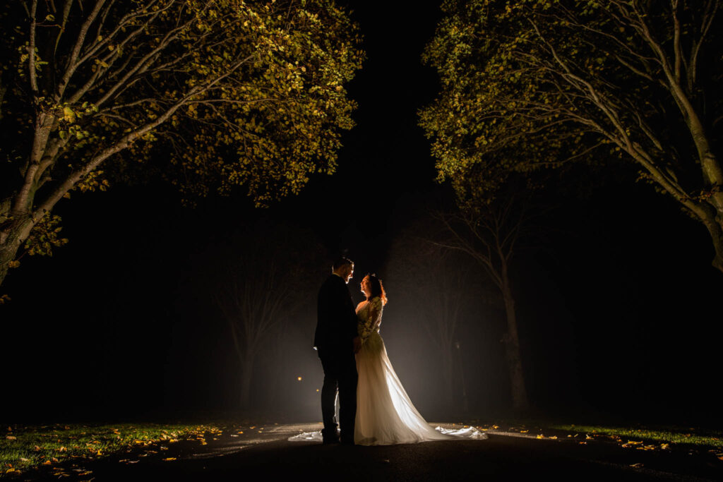 Bride and Groom silhouetted in from of a bright light as they stand facing each other standing under two over hanging trees